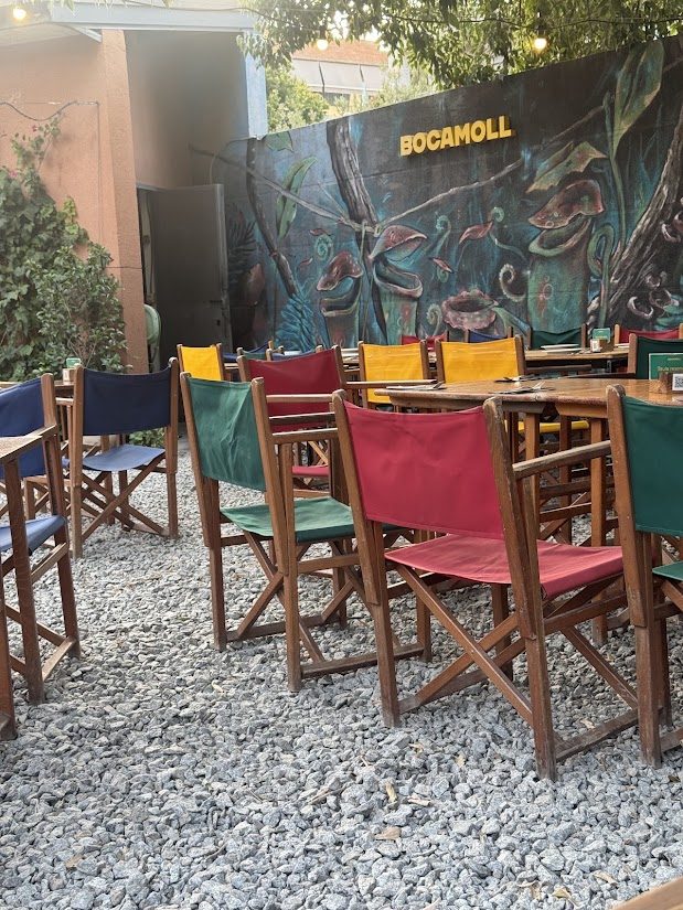 Terraza del Bar Bocamoll con las mesas de madera y las sillas de lona de colores delante del mural